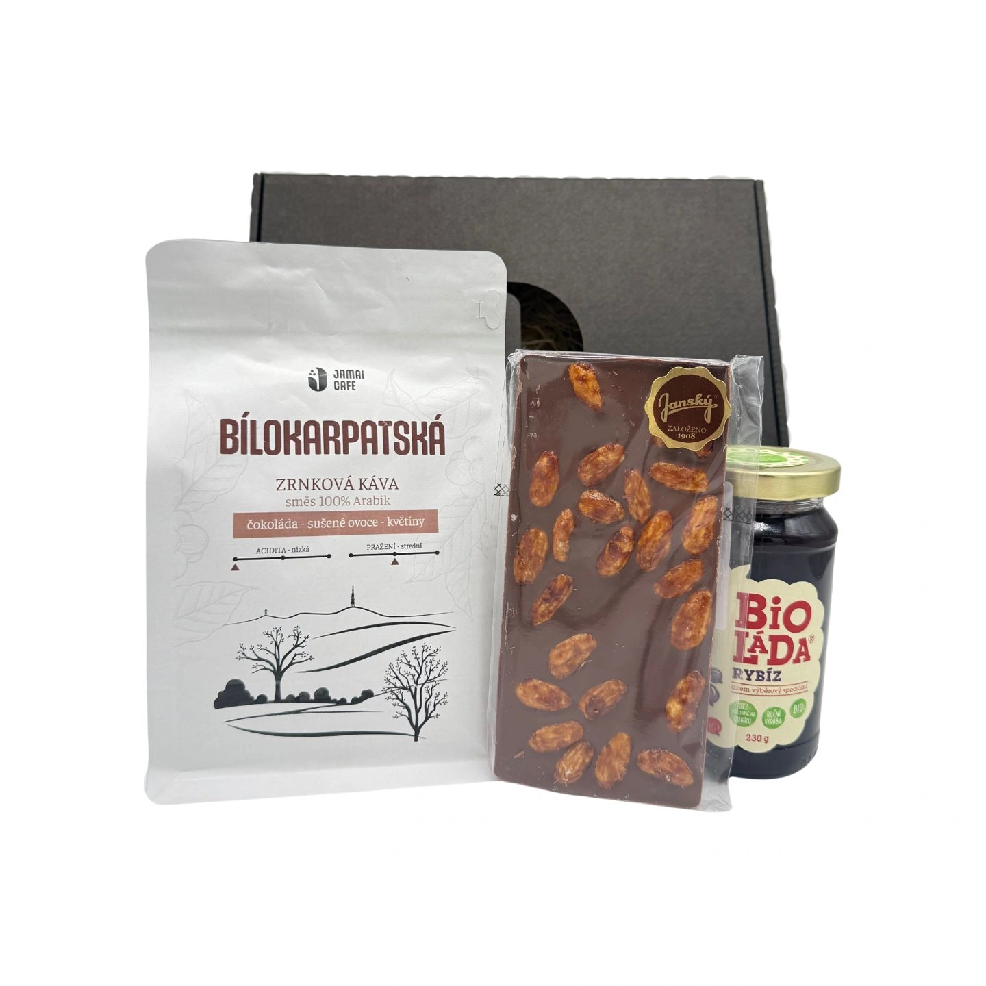 Dárkový balíček s kávou Bilokarpatská, tabulkou mléčné čokolády s ořechy a sklenicí rybízové Bio marmelády, vše před černou dárkovou krabicí.