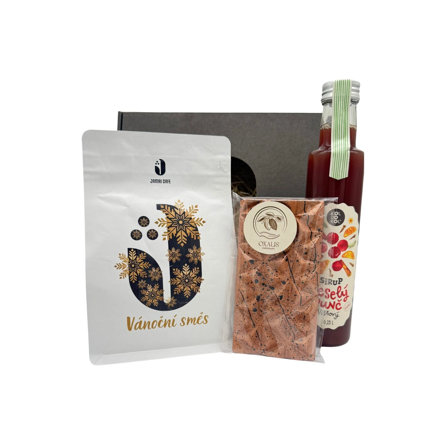 Dárkový balíček s kávou Vánoční směs, designovou čokoládou Oxalis a lahví ovocného sirupu Veselý punč vystavený před černou dárkovou krabicí.